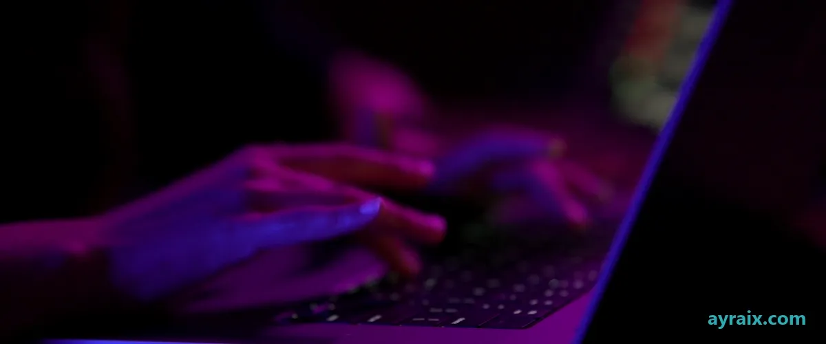 Hands typing on a laptop keyboard in a dark room, lit only by the screen in violet and magenta