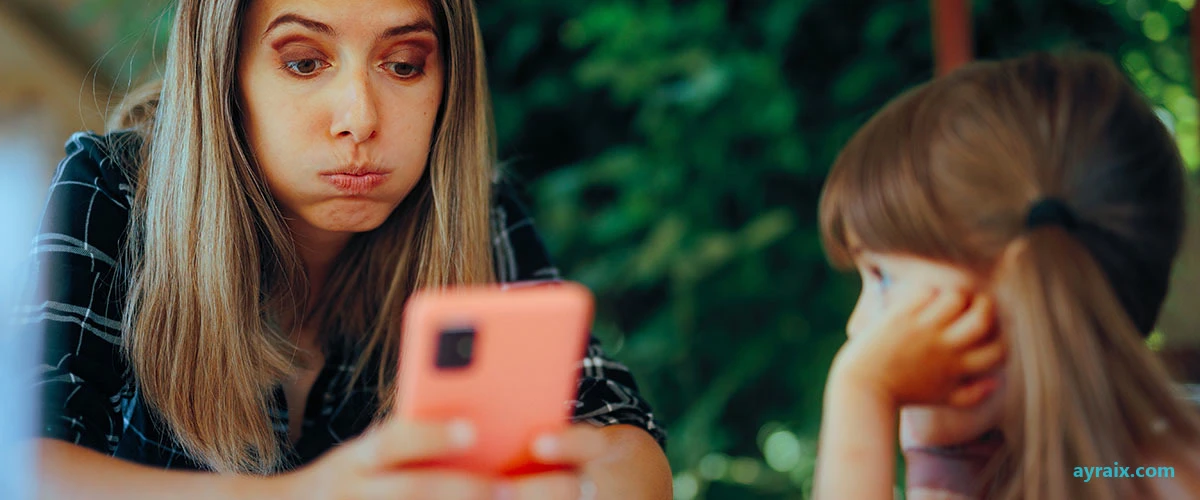 A mother checking her phone next to her daughter, both distracted by separate screens at the same table